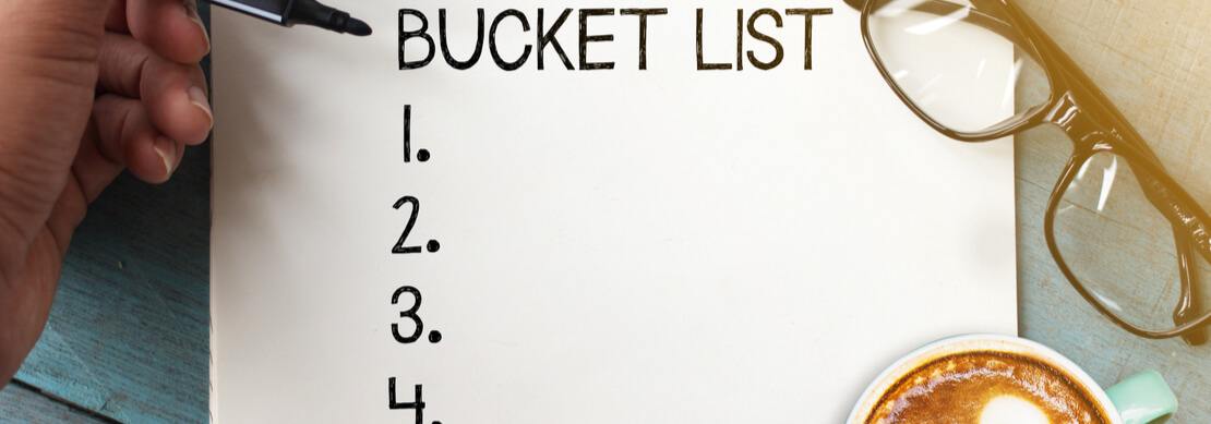 a hand writing a bucket list on a pad of paper with glasses and a cup of coffee on the side