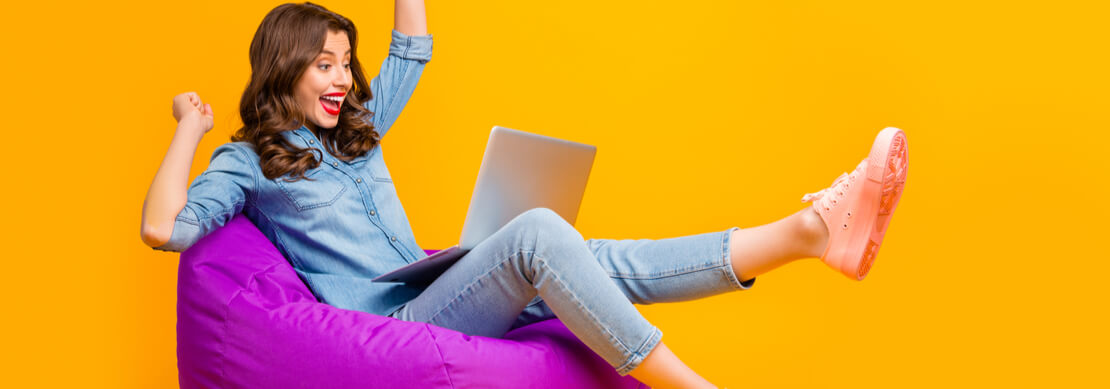 young woman sitting on a purple beanbag chair excited about winning on her laptop