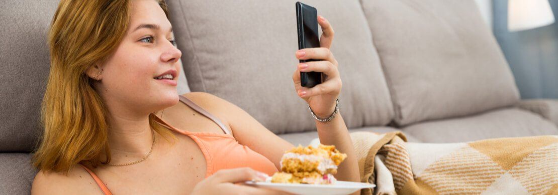 young woman laying on the sofa looking at his phone while eating
