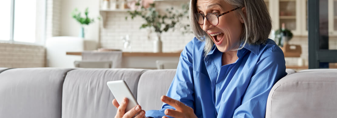 Older woman with long white hair happily playing casino games on mobile while comfortably sitting on her sofa