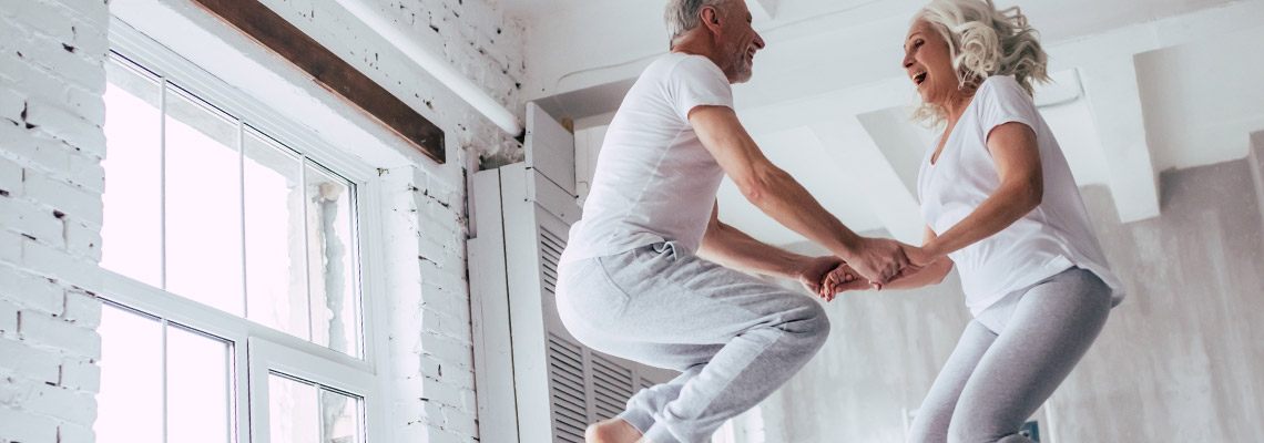 an older couple having fun jumping on their bed like little kids.  It is morning and the sun shines brightly in the window