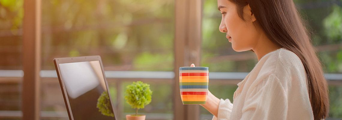 alert and attractive woman with long hair at the computer in a room with big windows and holding a cup of water