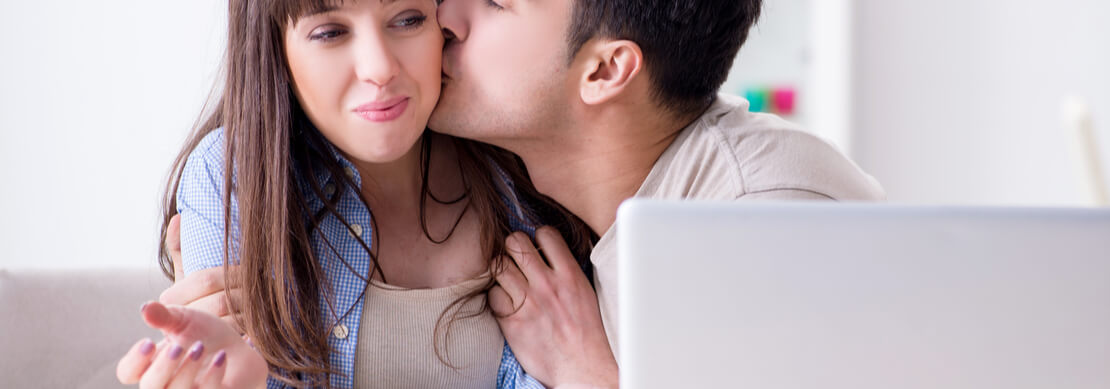 young couple playing at an online casino on their laptop. The man is kissing the women on the cheek and she has a bemused smile