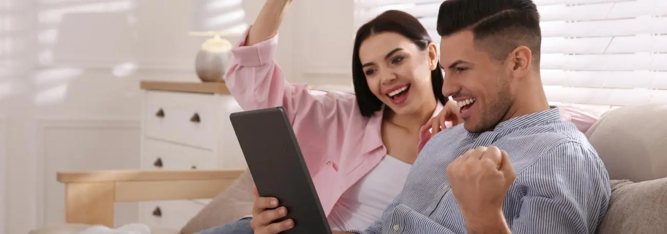 a young couple siing on the sofa with a tablet happily reacting to what they see which could be winning at an online casino