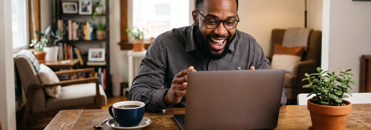 man in his 30's excitedly looking at his laptop with a small potted plant to his right and a cup of coffee to his left