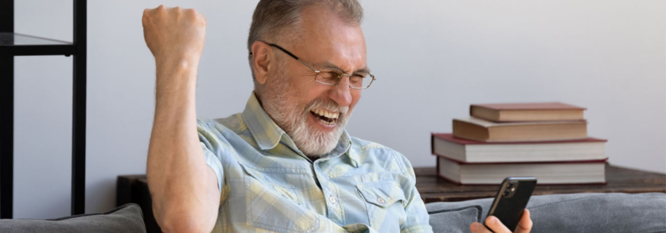 older man with short white beard happily enjoying online gaming on his smartphone. There are four books behind him on a table.
