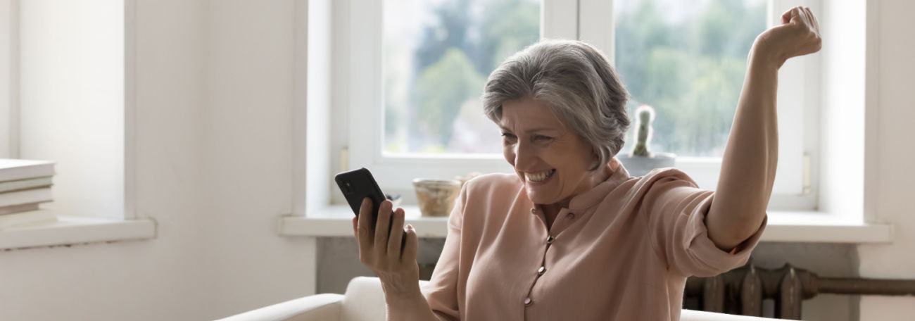 an older woman playing on a smartphone on the sofa ecstatically raising an arm after she wins a progressive jackpot