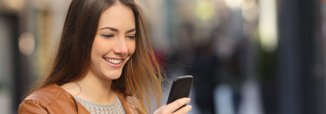 young woman outdoors with long brown hair smiles as she plays a calming casual game on her mobile device. 