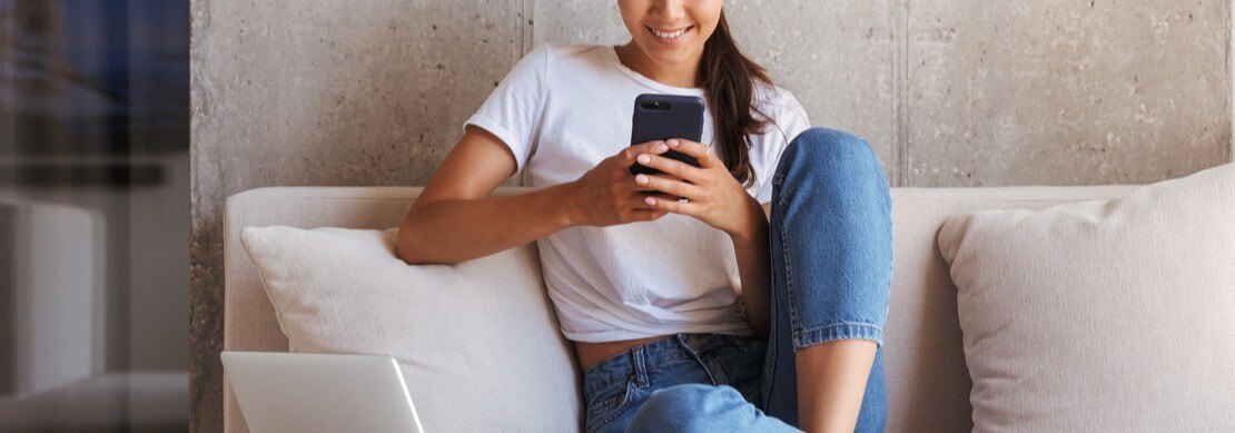young woman happily playing Jackpot Capital games on her phone sitting on her sofa