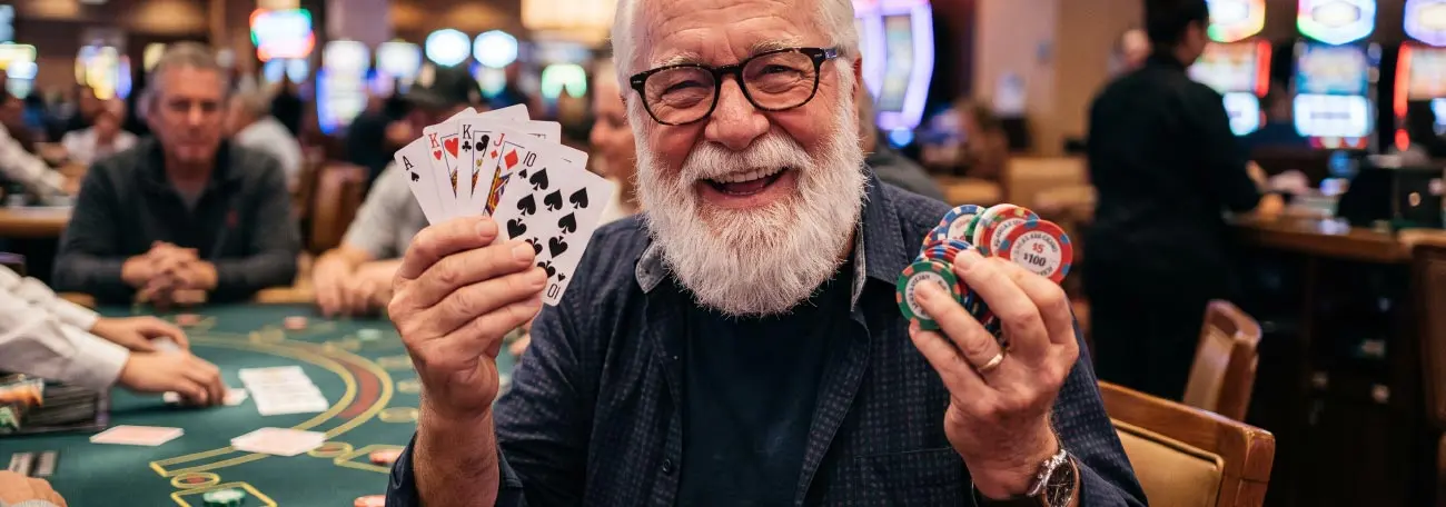 man with white beard and dark rimmed glasses smiling broadly holding playing cards in one hand and casino chips in the other