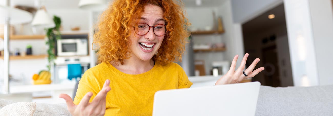 a happy young woman comfortably on her sofa with orange hair and a huge smile as she looks at her laptop computer