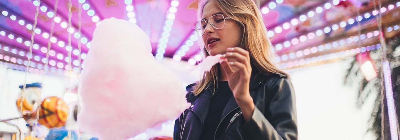 thirty-sh woman enjoying cotton candy at a local county fair. She is under a canopy of lights with fair rides behind her.