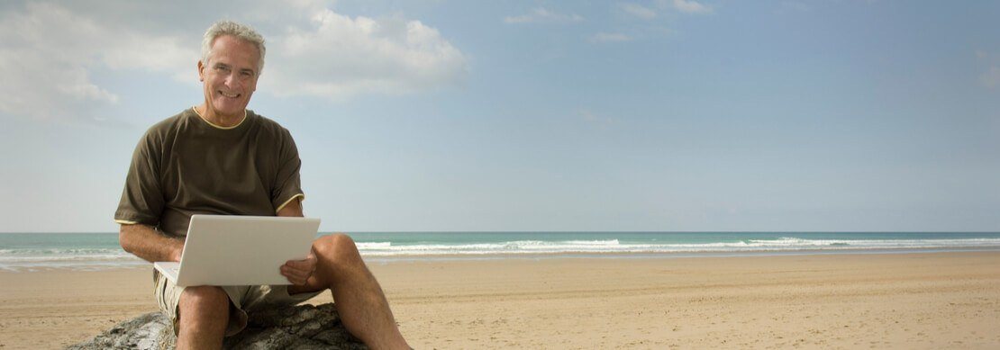 middle-aged man sitting on a stone on a beach playing on his laptop