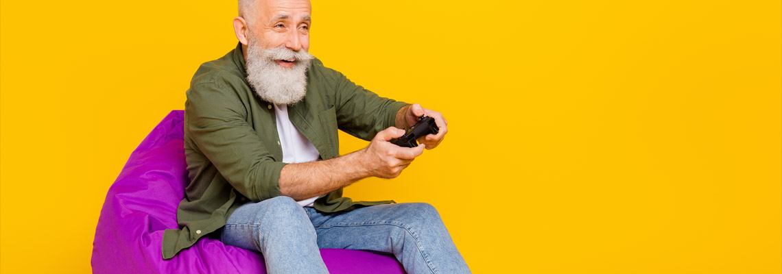 smiling elderly man with a long, white bread, sitting on a purple puff pillow with an open button down shirt playing games