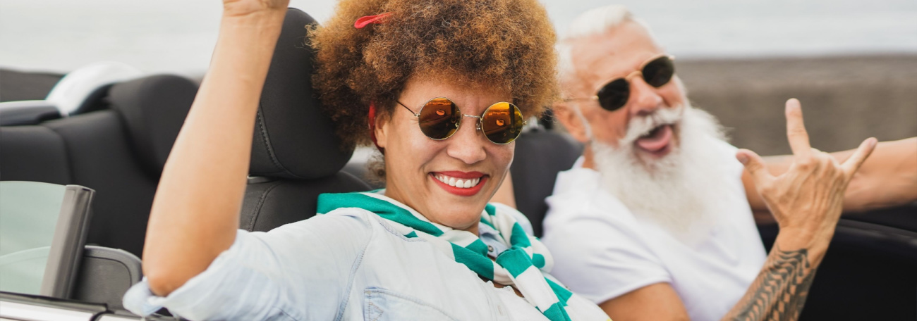 happy older couple in a convertible car. he has a white beard and she has an reddish orangeish afro.