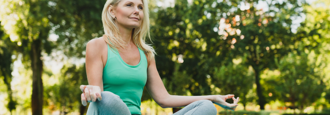 middle aged woman with long blond hair practicing meditation on a blanket in an open area of a park surrounded by trees