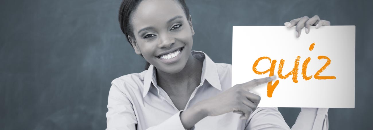 smiling young African American teacher sitting at her desk holding up a sign that says quiz.